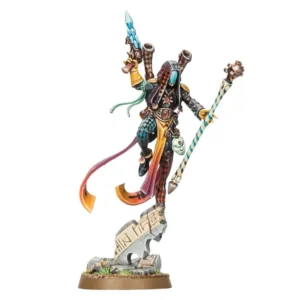 Photographie de studio sur fond blanc uni présentant la figurine officielle peinte d'un Prophète de l'Ombre, un sorcier mystique de la faction Aeldari pour l'univers Warhammer. Le personnage est représenté dans une pose acrobatique aérienne et dynamique, s'élançant avec légèreté depuis le sommet d'une ruine de pierre grise sculptée de runes. Il arbore une combinaison ajustée et une capuche ornées de motifs de losanges asymétriques noirs, rouges et bleus, complétées par un masque lisse turquoise miroitant sans aucun trait de visage. Deux lances-grenades dorsaux en forme de tubes cuivrés pointent derrière ses épaules, tandis que de longs rubans fluides jaune et orange ondulent dans son dos. De sa main droite levée, il brandit un pistolet à neuro-baisers noir surmonté d'un cristal bleu translucide, et de sa main gauche, il tient un long bâton de brume peint de rayures spirales blanches, bleues et jaunes, couronné d'un crâne stylisé en métal cuivré. Un second masque rituel blanc miniature est suspendu à sa ceinture. La figurine repose sur un socle rond texturé de terre marron et de petites touches d'herbe verte.