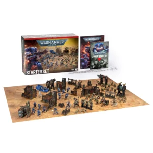 A wide, overhead product shot displaying the full contents of the Warhammer 40,000: Starter Set (Anglais), presented against a solid white background. In the upper left, the official box stands upright, featuring a red top banner reading "START HERE", the silver "WARHAMMER 40,000" logo, and the bold text "STARTER SET" at the bottom over an illustration of a blue Space Marine in battle. To its right are two booklets, including a "CORE RULES" guide showing a blue helmet on its cover, a small pile of white six-sided dice, and transparent plastic range rulers. Spanned across the lower half is a large, sandy-colored gaming mat depicting a desert battlefield crowded with fully painted miniatures and scenery. On the left side, the blue-armored Space Marines stand ground near dark, multi-level ruined gothic buildings and a blue floating vehicle. On the right, a large horde of green-skinned Orks wielding axes, firearms, and ramshackle scrap-metal vehicles charges through matching mechanical barricades and towers, creating an immersive battlefield scene.