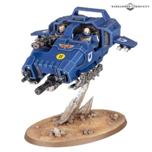 Photo de présentation sur fond blanc d'un véhicule antigrav Land Speeder des Space Marines, peint aux couleurs bleues et dorées du chapitre des Ultramarines. L'appareil blindé présente une carrosserie bleue ornée du symbole blanc « U » de la faction et d'un marquage jaune avec le chiffre romain « II » sur la proue. Dans le cockpit ouvert, deux Space Marines en armure sont visibles : le pilote aux commandes et un artilleur à ses côtés. L'armement comprend un canon d'assaut rotatif monté sous le nez, un bolter lourd frontal et des missiles fixés sous les ailes latérales. Le véhicule est en position de vol au-dessus d'un socle d'exposition sablonneux décoré de rochers et d'un crâne, maintenu par un effet sculpté de fumée grise. Le logo « WARHAMMER COMMUNITY » figure en haut à droite.